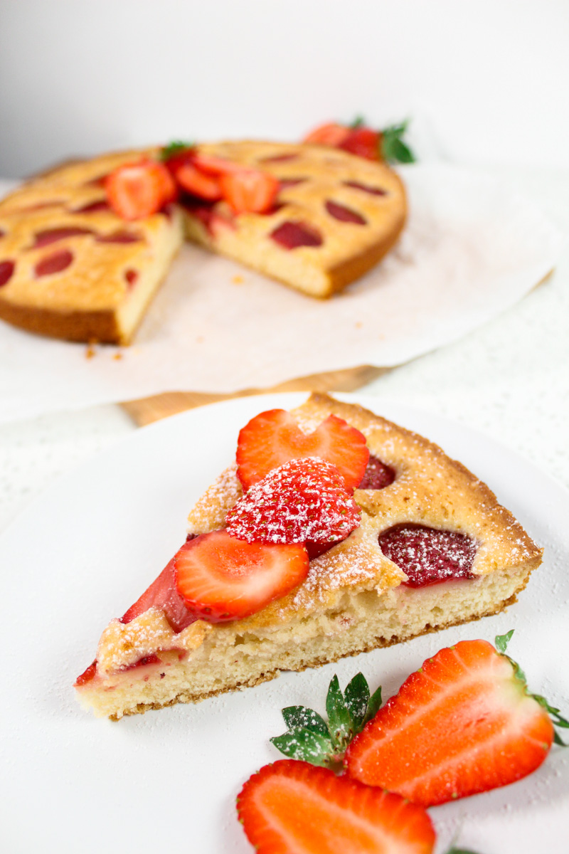 A piece of strawberry cake in a plate with two strawberries in front of it and the rest of the cake in the background